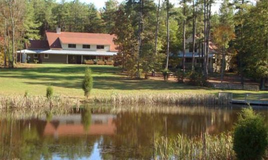 Camp Royall can be seen from across the still waters of a small pond on a sunny day.